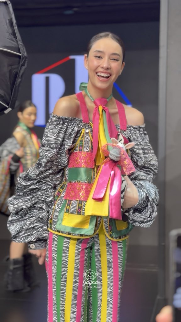 A beaming model at Siam Center BIFW26 showcases a vibrant Ginjay ensemble featuring a zebra-print off-shoulder top paired with colorful striped trousers and an oversized ribbon neckpiece, holding a plush bunny accessory. The image, tagged with #SiamCenterBIFW2026xGINJAY, captures the playful spirit of the fashion event.