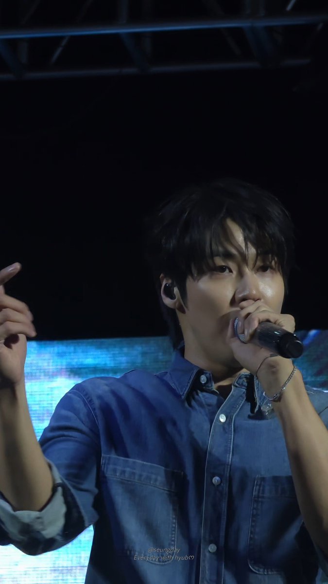 A male performer singing into a microphone on stage while wearing a blue button-up shirt and an earpiece.