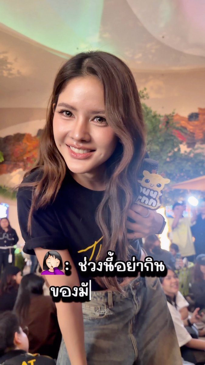 A young woman with long wavy brown hair smiles at the camera while holding a microphone with a cute character logo; overlaid Thai text reads 'ช่วงนี้อย่าถ่ายของผี' with an emoji of a woman covering her face.