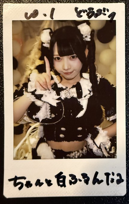 A Polaroid-style photo shows a young woman with dark hair in pigtails wearing a black and white frilly maid-style outfit, posing with her index finger raised; Japanese text is handwritten on the border.