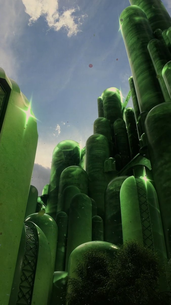 Tall, glowing green cylindrical pods rise like crystalline towers toward a bright blue sky, evoking the shimmering emerald City of Oz from Wicked, perfectly matching the "GLINDA GLITTER #WOZatSphere" post celebrating the immersive experience at the Sphere.