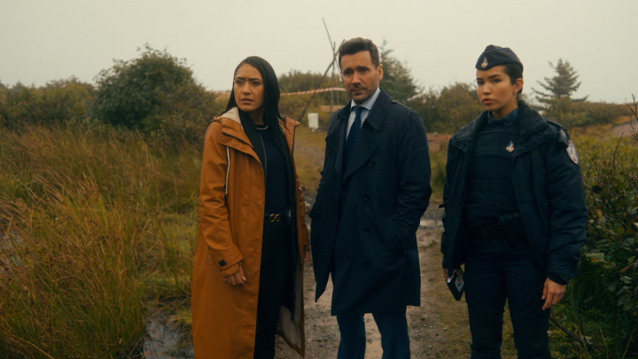Three people—a woman in a brown coat, a man in a dark suit, and a woman in a police uniform—standing in a grassy, overcast outdoor setting that appears to be a crime scene.