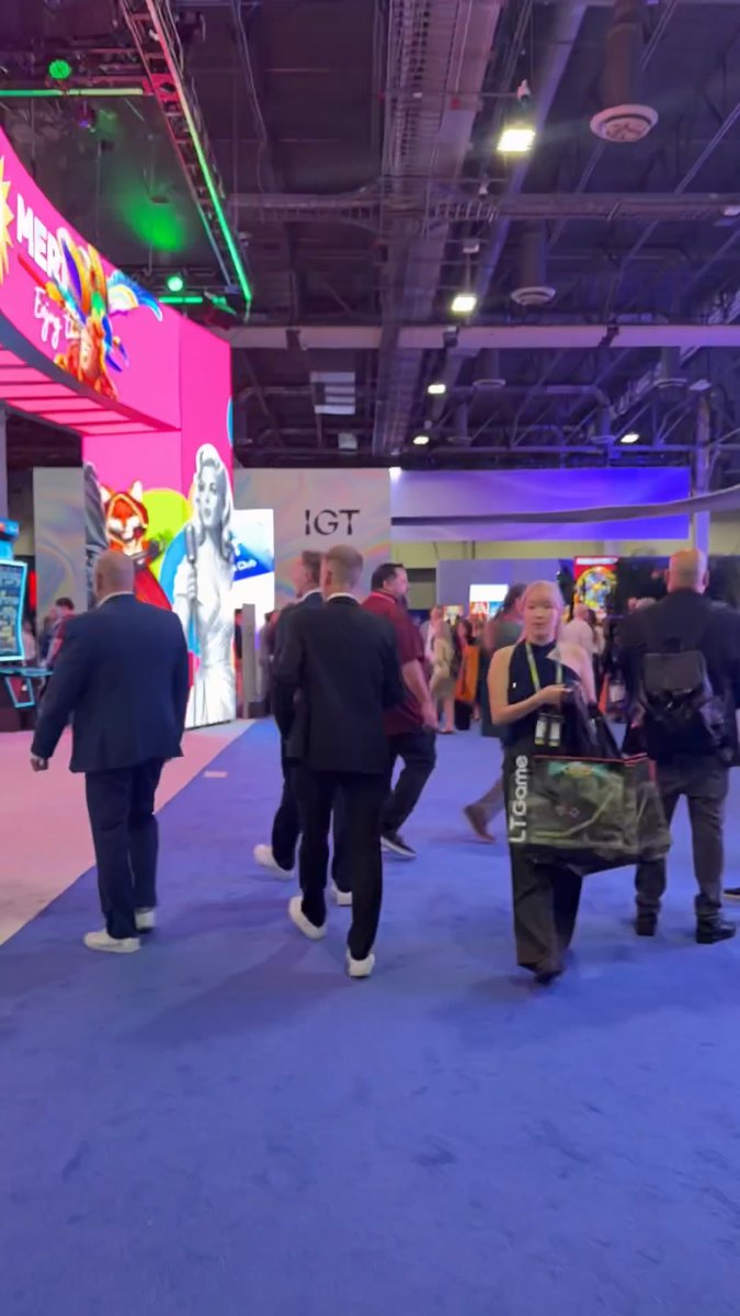 A bustling trade show floor with people walking on a blue carpeted area, featuring colorful booth displays including a bright pink booth and a sign reading 'IGT' in the background.