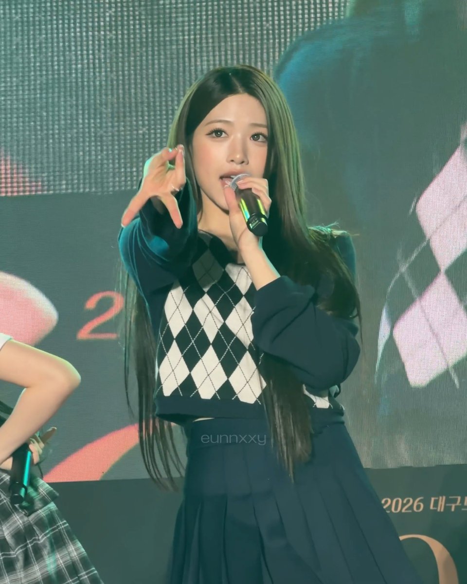 A K-pop idol with long dark hair performing on stage with a microphone, wearing a black and white argyle sweater; text on screen includes 'eunnxxy' and '2026 대구'.