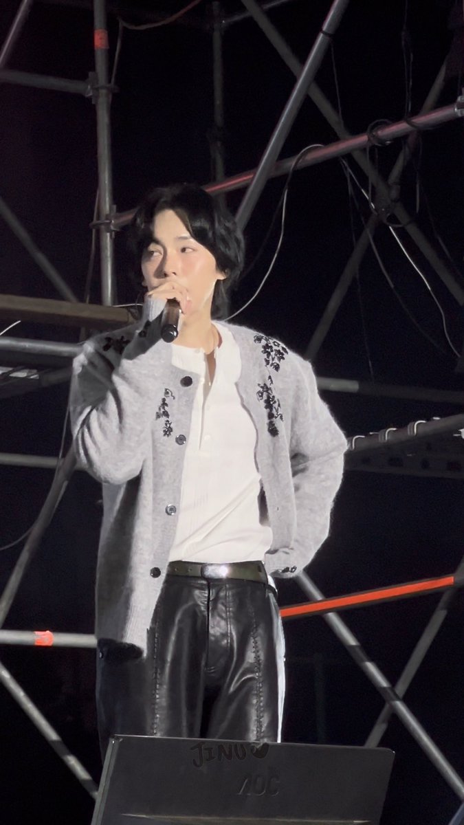 Suga (Min Yoongi) of BTS performing on stage, singing into a microphone while wearing a grey cardigan over a white shirt and black leather pants, with 'JINO ZOC' visible on his laptop.