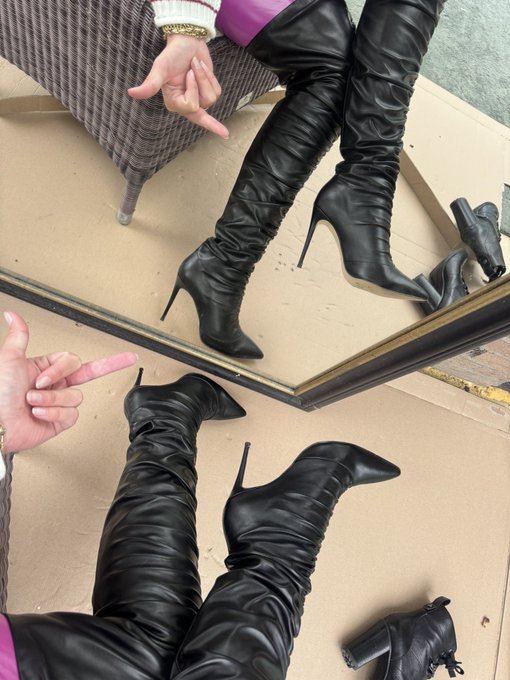 A mirror selfie showcasing black thigh-high stiletto boots paired with a purple garment, with the person pointing at their reflection.