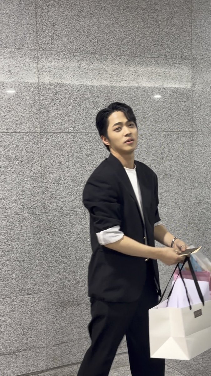 A young man wearing a black blazer over a white t-shirt walks past a grey stone wall, carrying a white shopping bag and some items.