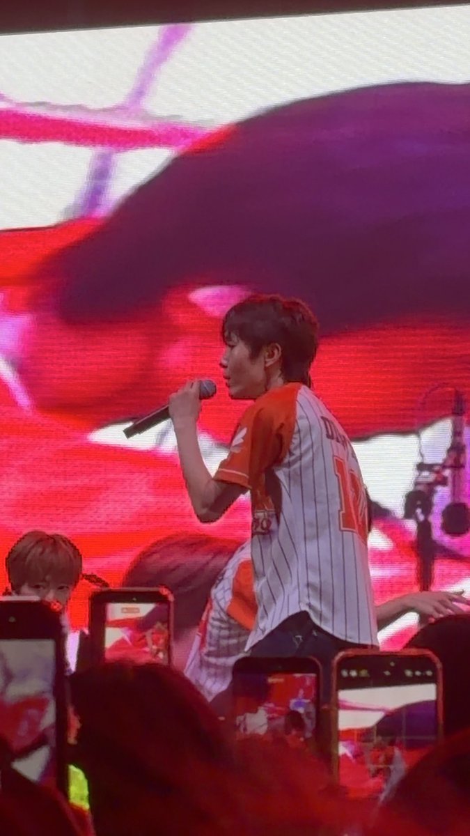 A concert photo showing a performer in a red and white striped jersey singing into a microphone on stage with a large red-lit screen backdrop, while audience members record with phones.