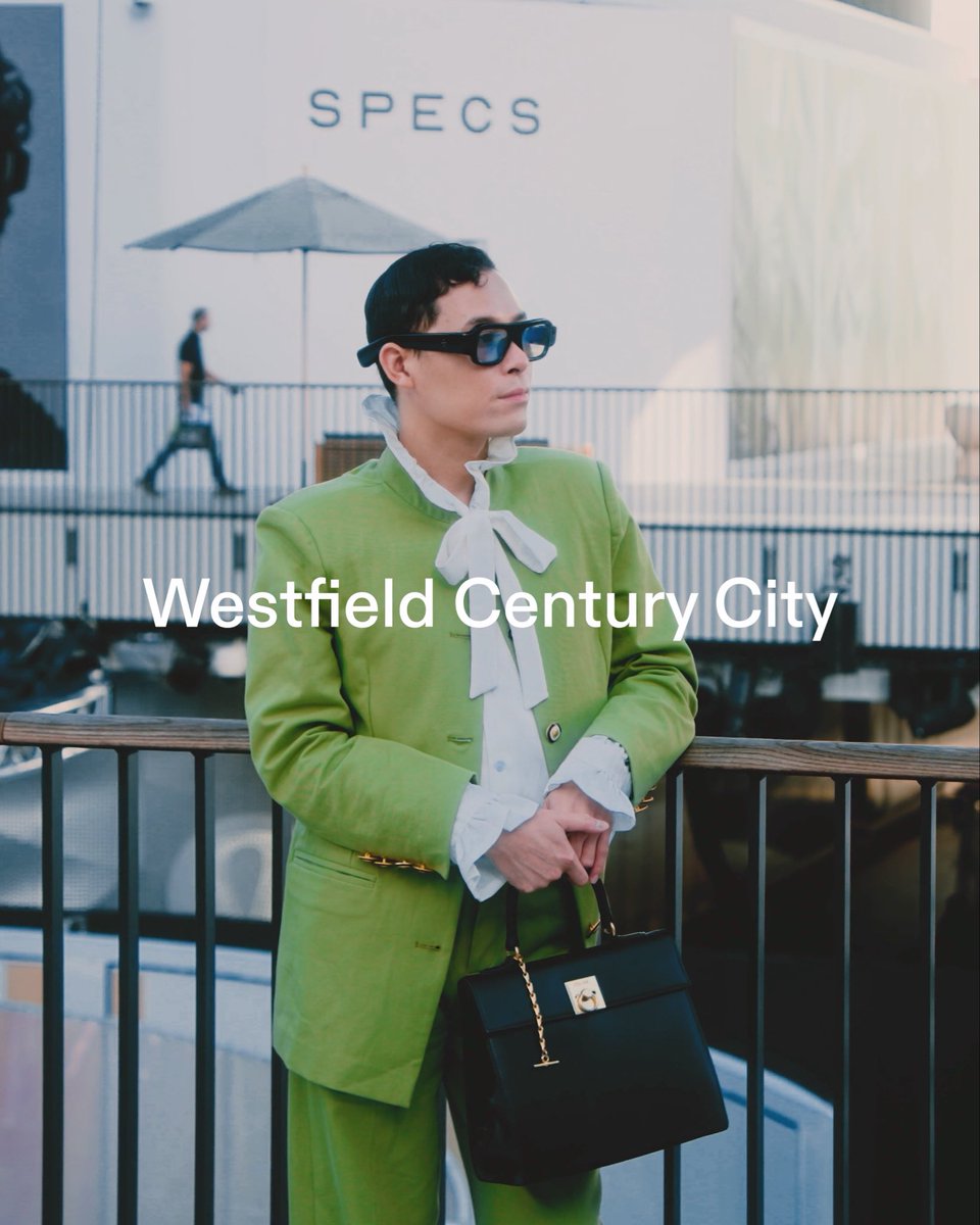 A man wearing a green suit, white ruffled blouse, and black sunglasses stands holding a black handbag, with text reading 'SPECS' and 'Westfield Century City' overlaid on the image.