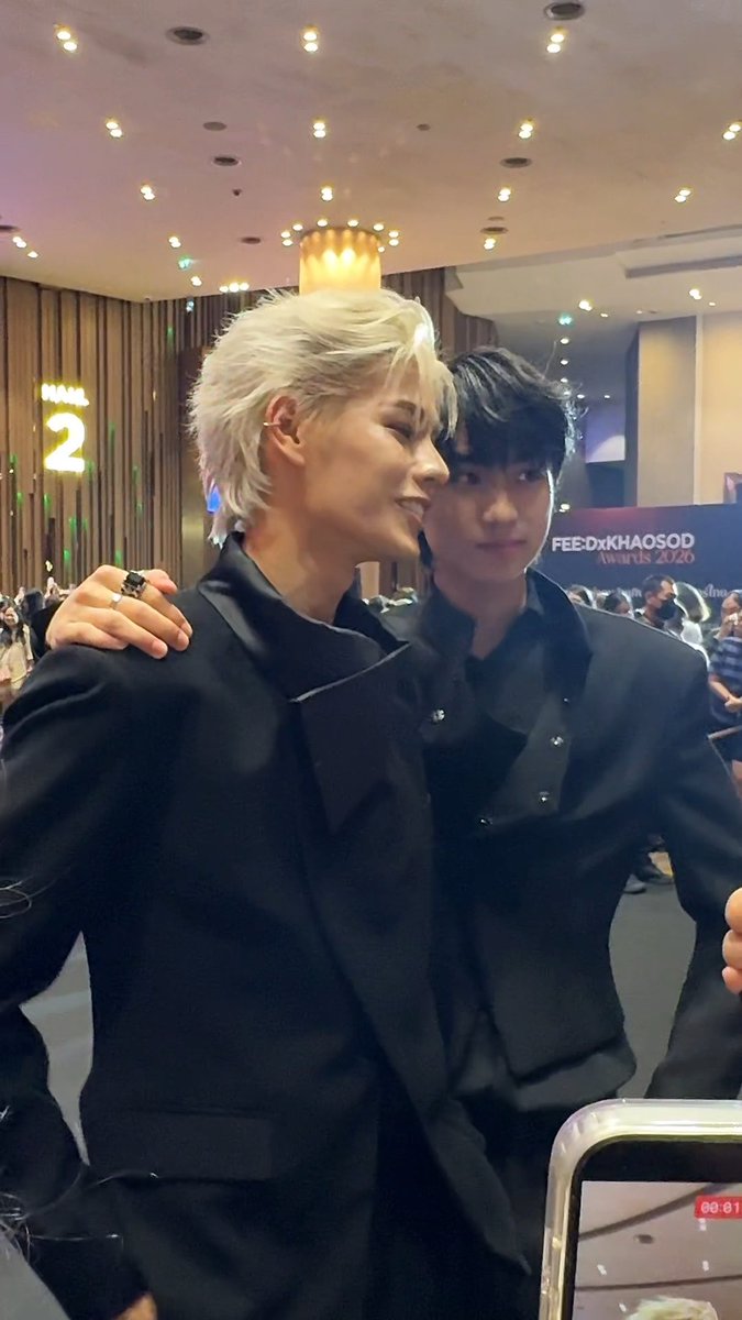Two young men in black suits pose at an event; the blonde one has his arm around the dark-haired one, with 'FEE Dx KHADSOD Awards 2026' and '2' visible in the background.