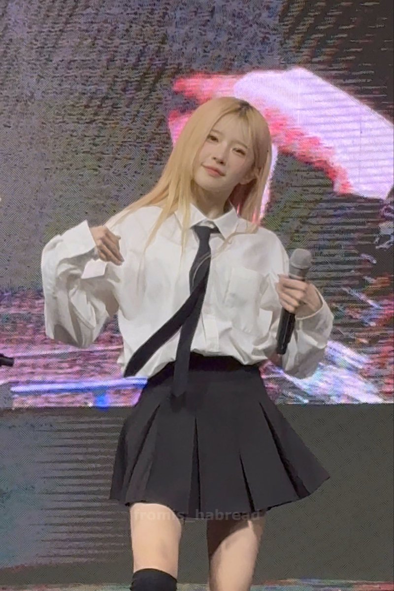 A blonde K-pop idol performing on stage in a school uniform outfit consisting of a white shirt, black tie, and black pleated skirt, with a 'from: habread' watermark.