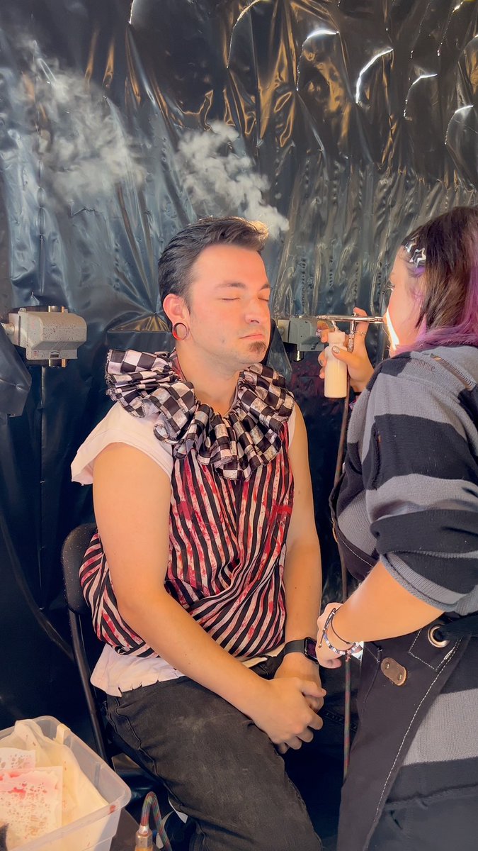 A man in a striped apron and ruffled clown collar sits with his eyes closed while a person with purple-streaked hair uses a spray bottle and tool near his face, with visible smoke in the background.