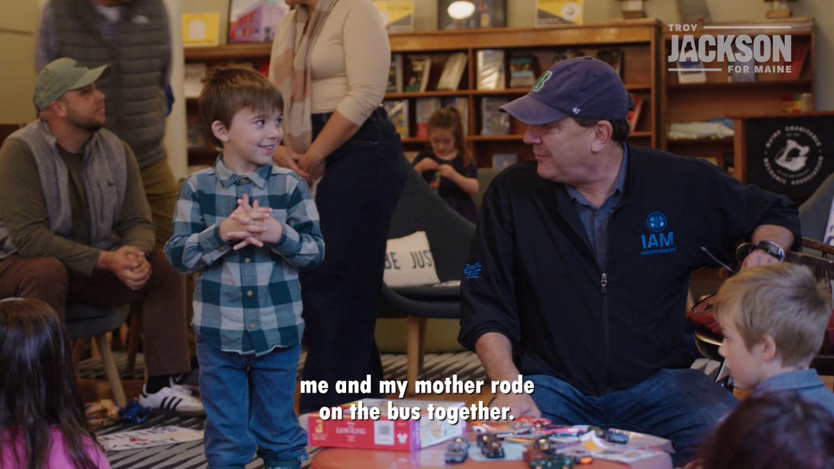 A man identified as Troy Jackson, in a cap and dark "IAM" jacket, kneels to listen to a smiling young boy sharing a personal story in what appears to be a bookstore or community space, with the caption "me and my mother rode on the bus together." The image accompanies a post criticizing tax breaks for the wealthy at the expense of child welfare, illustrating Jackson's connection to everyday Maine families and children.