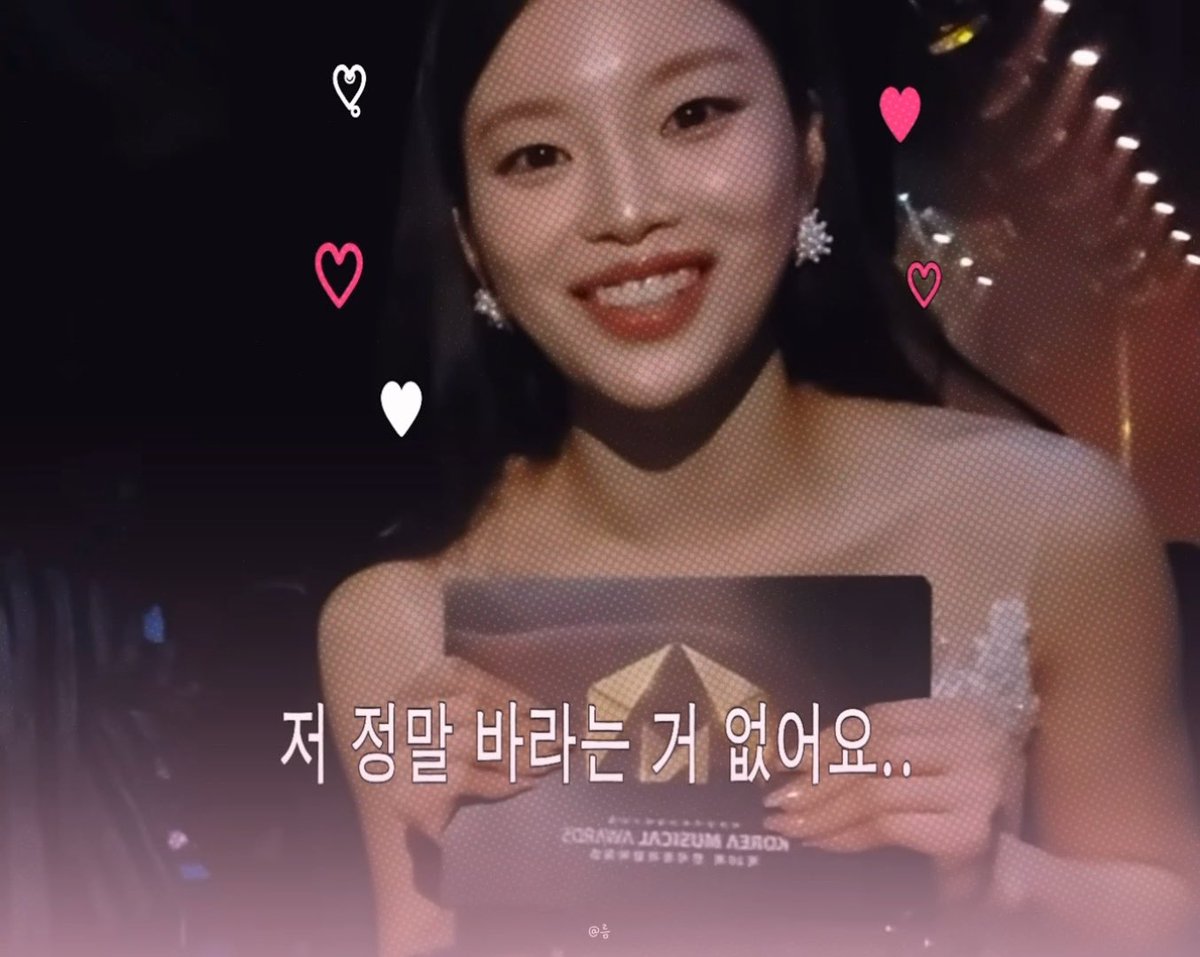 A young woman with long dark hair, wearing a white off-shoulder dress and sparkly earrings, smiles brightly while holding a "KOREA MUSIC AWARDS" trophy. The Korean text "저 정말 바라는 거 없어요.." (I don't really want anything..) appears at the bottom, surrounded by decorative heart graphics.