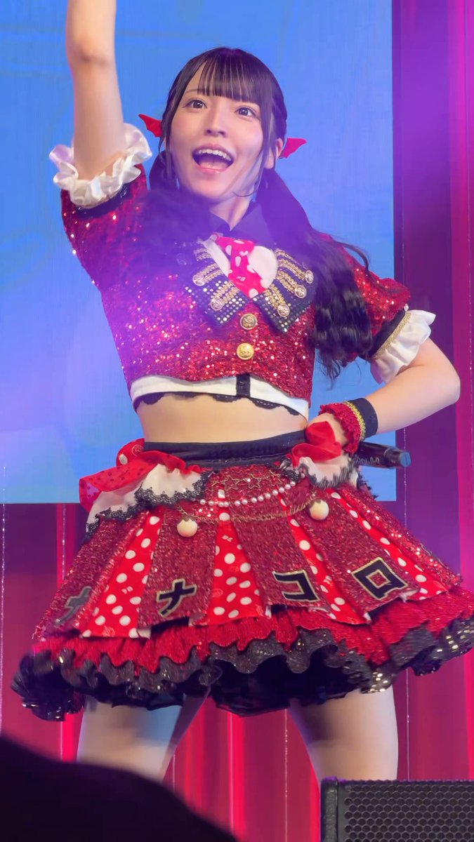 A young woman performs on stage in a red and black idol outfit with a sequined cropped vest, polka-dot skirt, and red hair bows, raising one arm while holding a microphone.