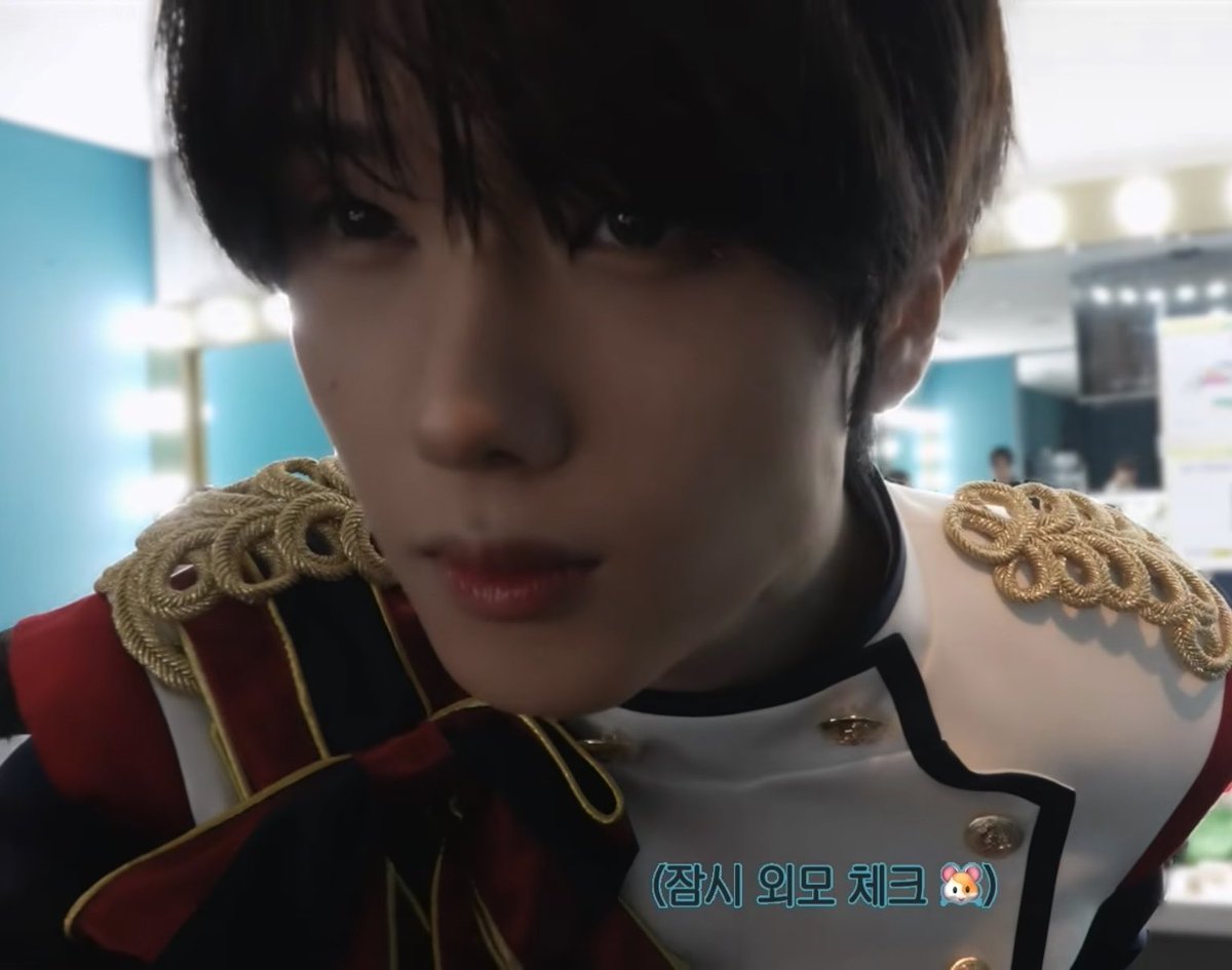 A close-up selfie of a young man with dark hair wearing an ornate white uniform featuring gold braided epaulettes, with Korean text overlay reading '(잠시 외모 체크' accompanied by a bunny emoji.