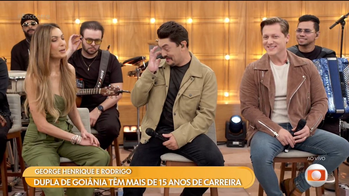 A TV broadcast scene featuring a woman in a green outfit, a man with a mustache in a tan jacket, and a smiling man in a brown leather jacket, with the caption 'GEORGE HENRIQUE E RODRIGO - DUPLA DE GOIÂNIA TÊM MAIS DE 15 ANOS DE CARREIRA'.