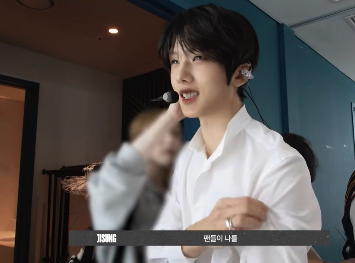 A young man with dark hair wearing a white button-down shirt and an earpiece microphone, smiling, with a caption identifying him as 'JISUNG' and Korean text below.