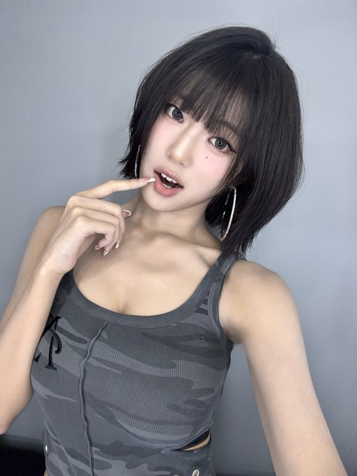 A young woman with short, layered black hair and large hoop earrings poses playfully, touching her lip, while dressed in a grey camouflage-patterned tank top.