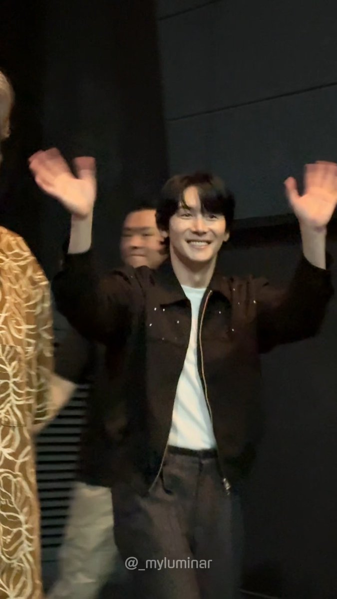 A man in a brown jacket and white shirt smiles broadly while raising both hands in a cheerful wave during what appears to be a stage greeting. The watermark "@_myluminar" is visible at the bottom, and the Korean caption confirms this is Kim Jun-han greeting fans at the curtain call for the play "Intern" (인턴) on September 27, 2026.