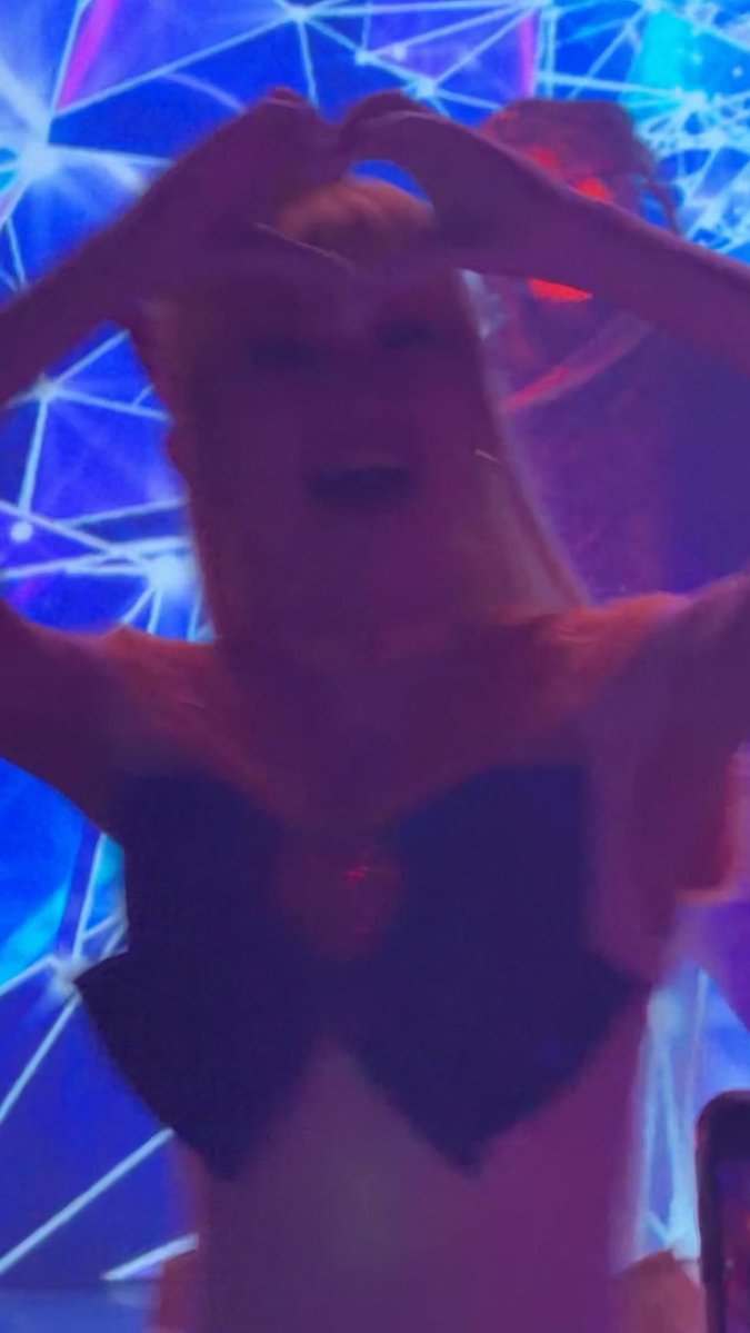 A woman in a black top poses with her hands forming a heart shape above her head, set against a blue neon-lit background with geometric patterns.