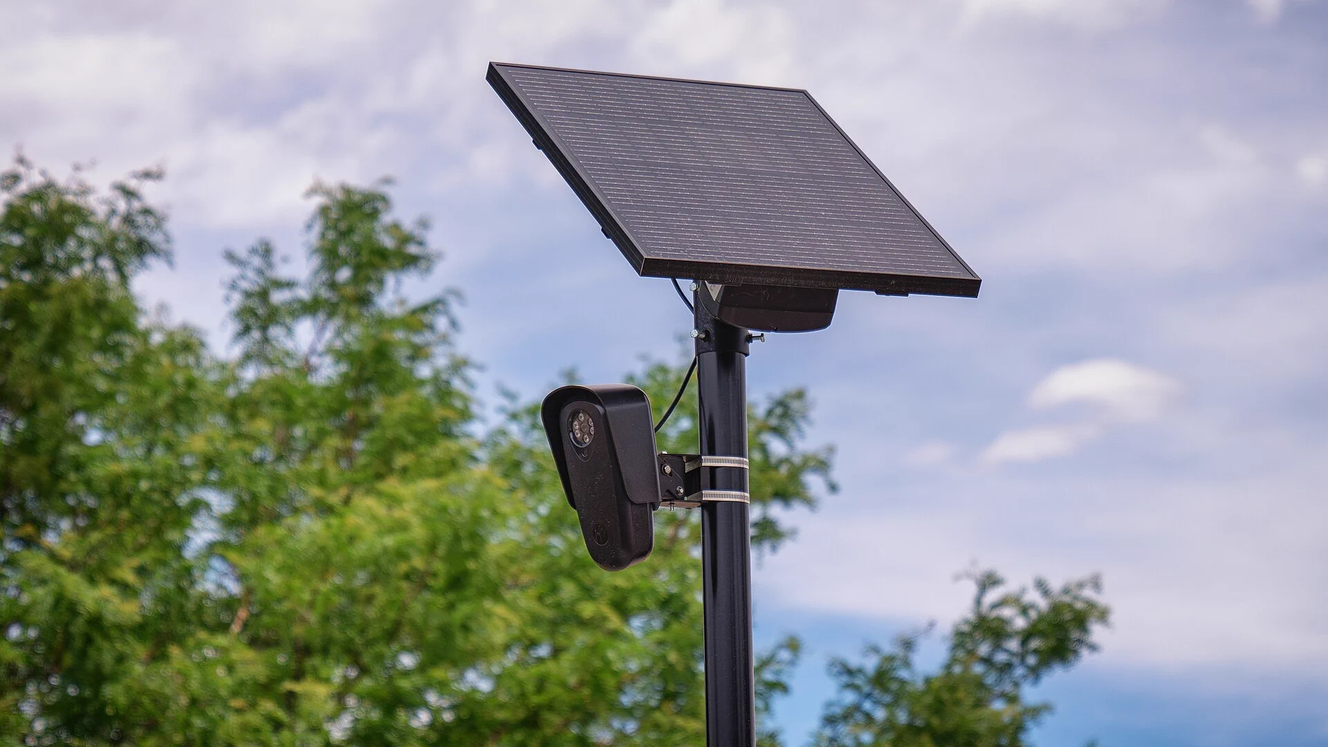 An image of a black flock camera outdoors. The camera is mounted on a black pole, and has a black, square solar panel above it. In the background, the sky is blue and cloudy, and the top of a tree with green leaves is visible.