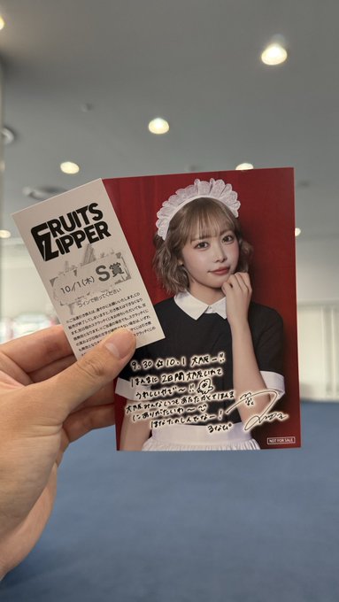 A hand holds up a handwritten 'FRUITS ZIPPER' photo card featuring a member in a maid outfit, dated 10/1(水) S賞, with a personal message addressed to 'てるこ' and signed 310(い).