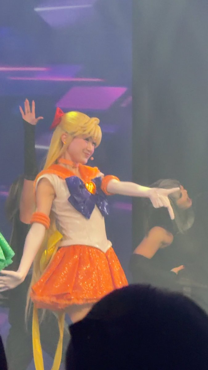 A performer on stage dressed as Sailor Venus from Sailor Moon, wearing the iconic orange skirt and blue bow, with blonde hair and a red bow accessory, posing with outstretched arm.