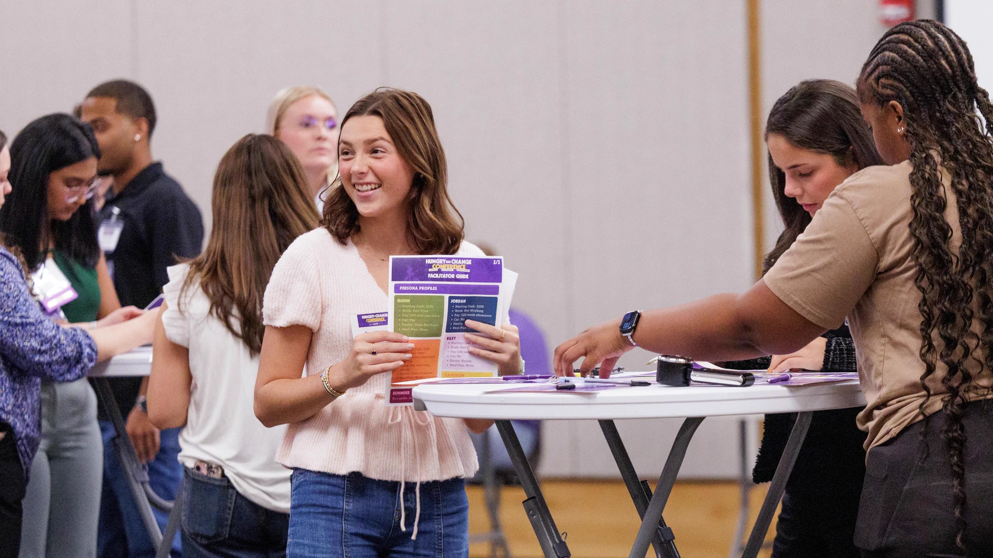 A woman smiles while holding a 'HUNGRY CHANGE CONFERENCE FACILITATOR GUIDE' pamphlet. Others are gathered around tables in a bright room, looking at materials.