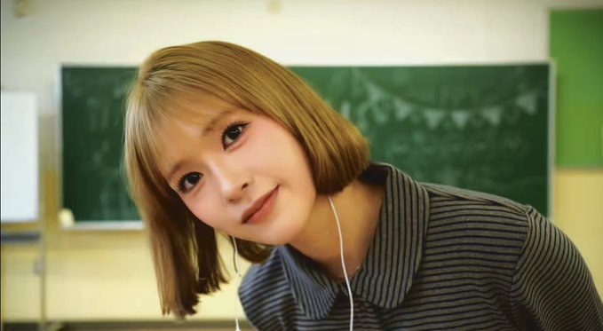 A young woman with short brown hair and earphones smiles while leaning forward in what appears to be a classroom setting.