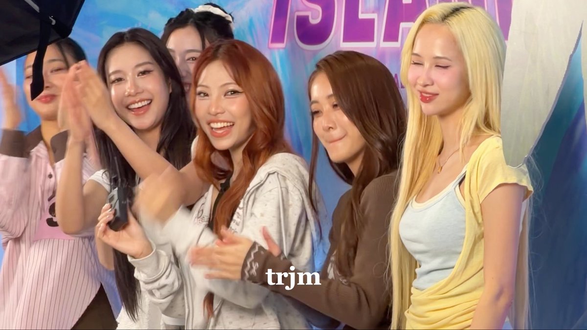 Five young women smile and pose together in a brightly lit room with blue walls and partial 'ISLAN' text visible in the background. The watermark 'trjm' appears on the image.