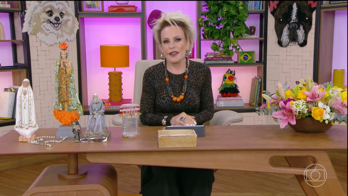 A blonde woman in a leopard-print top sits behind a wooden desk on a television set decorated with religious figurines, dog portraits, and a large floral arrangement.