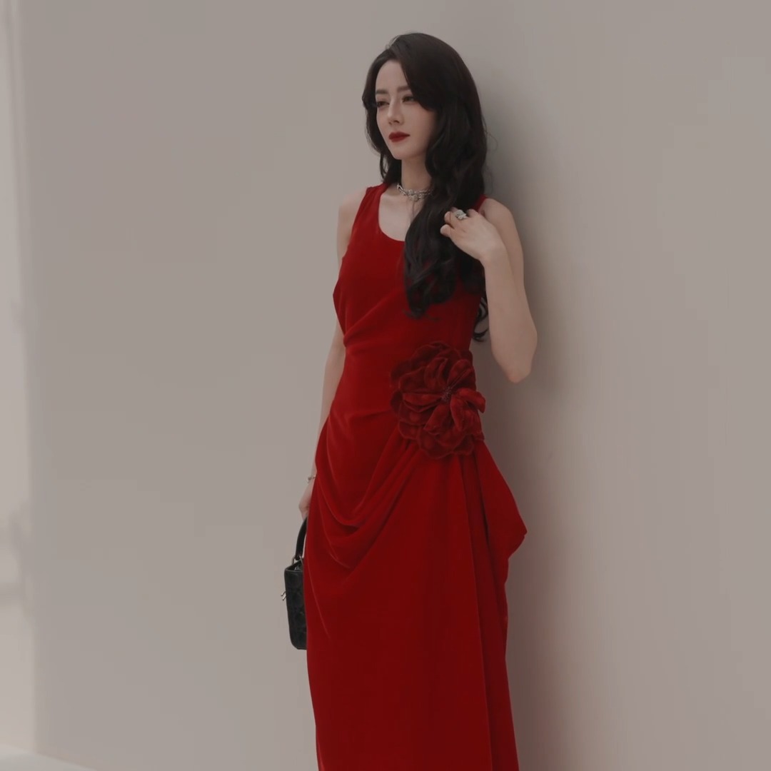 A stylish portrait of an Asian woman wearing an elegant, draped red sleeveless dress with a large floral detail, standing against a white wall.