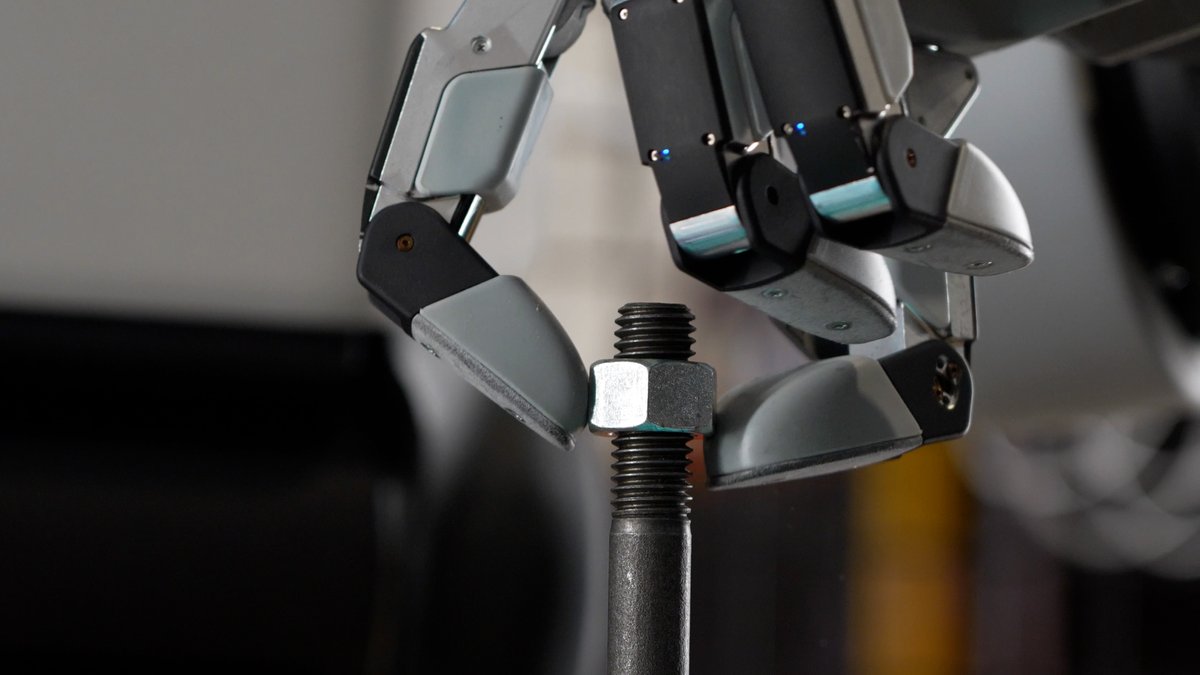 A robotic hand with gray fingers grips a metal nut threaded onto a vertical black rod.