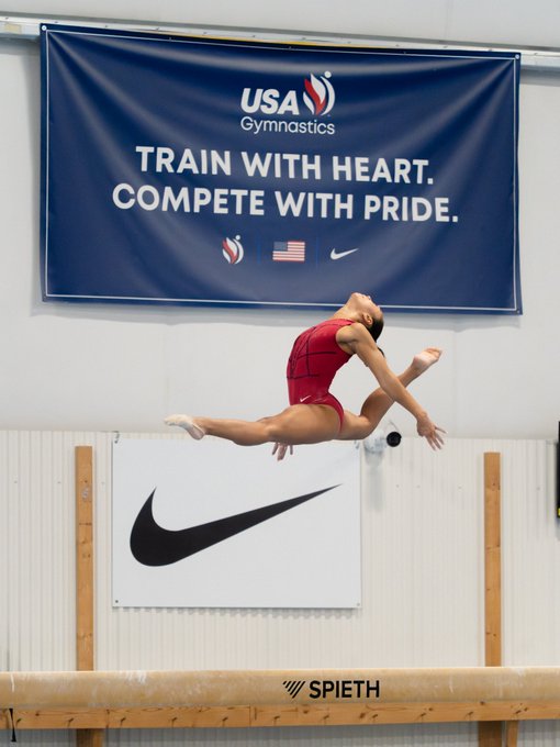 A gymnast in mid-air performs a leap on the balance beam during the Women's Worlds Selection Event, with a "USA Gymnastics" banner reading "TRAIN WITH HEART. COMPETE WITH PRIDE." hanging above and "SPIETH" branding on the beam below. The image captures the high-flying action from day two of competition.