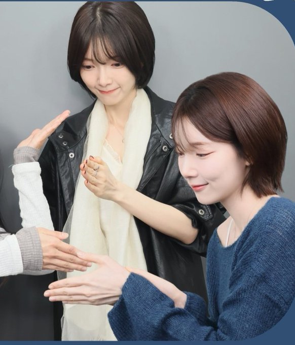 Two young women with short hair smiling and looking at their hands, with one wearing a black leather jacket and white scarf, and the other in a blue knit sweater, while someone else's hands reach in from the left.