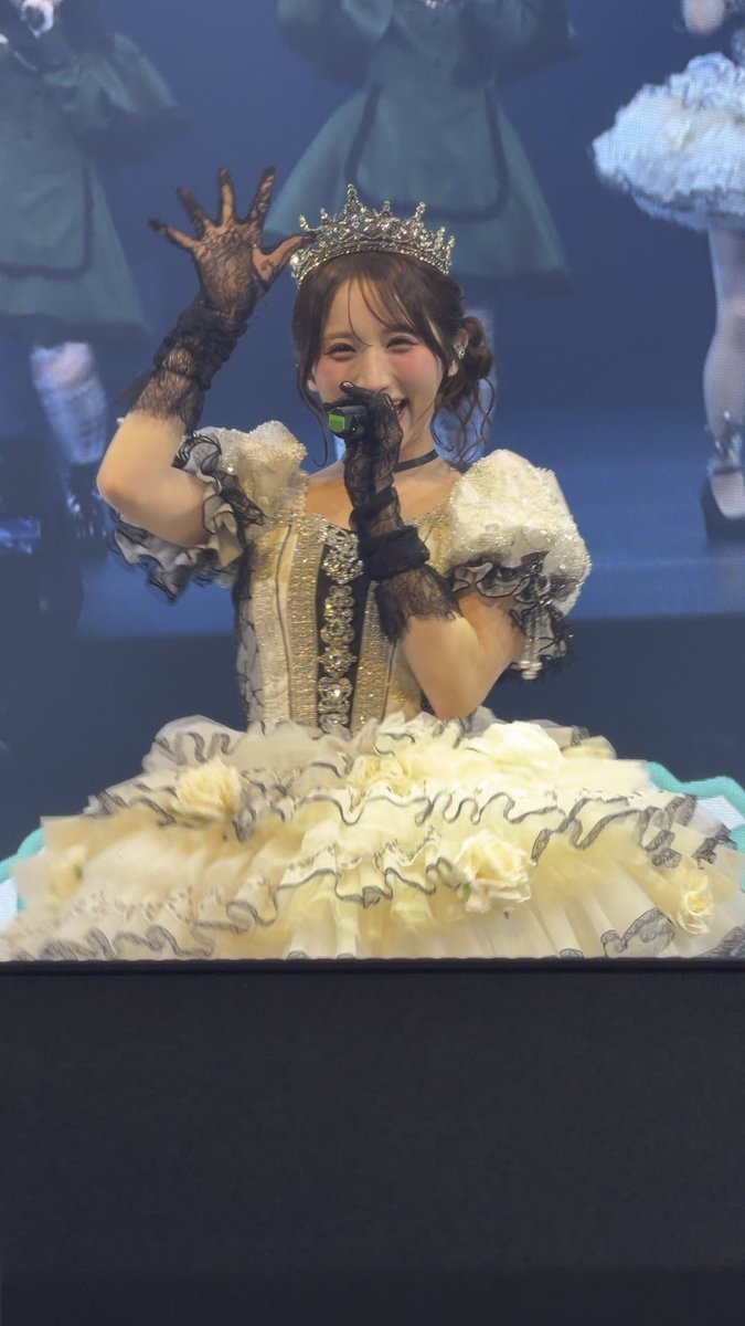 A young woman wearing an ornate white and black dress with a crown and black lace gloves smiles and waves while holding a microphone on a stage.