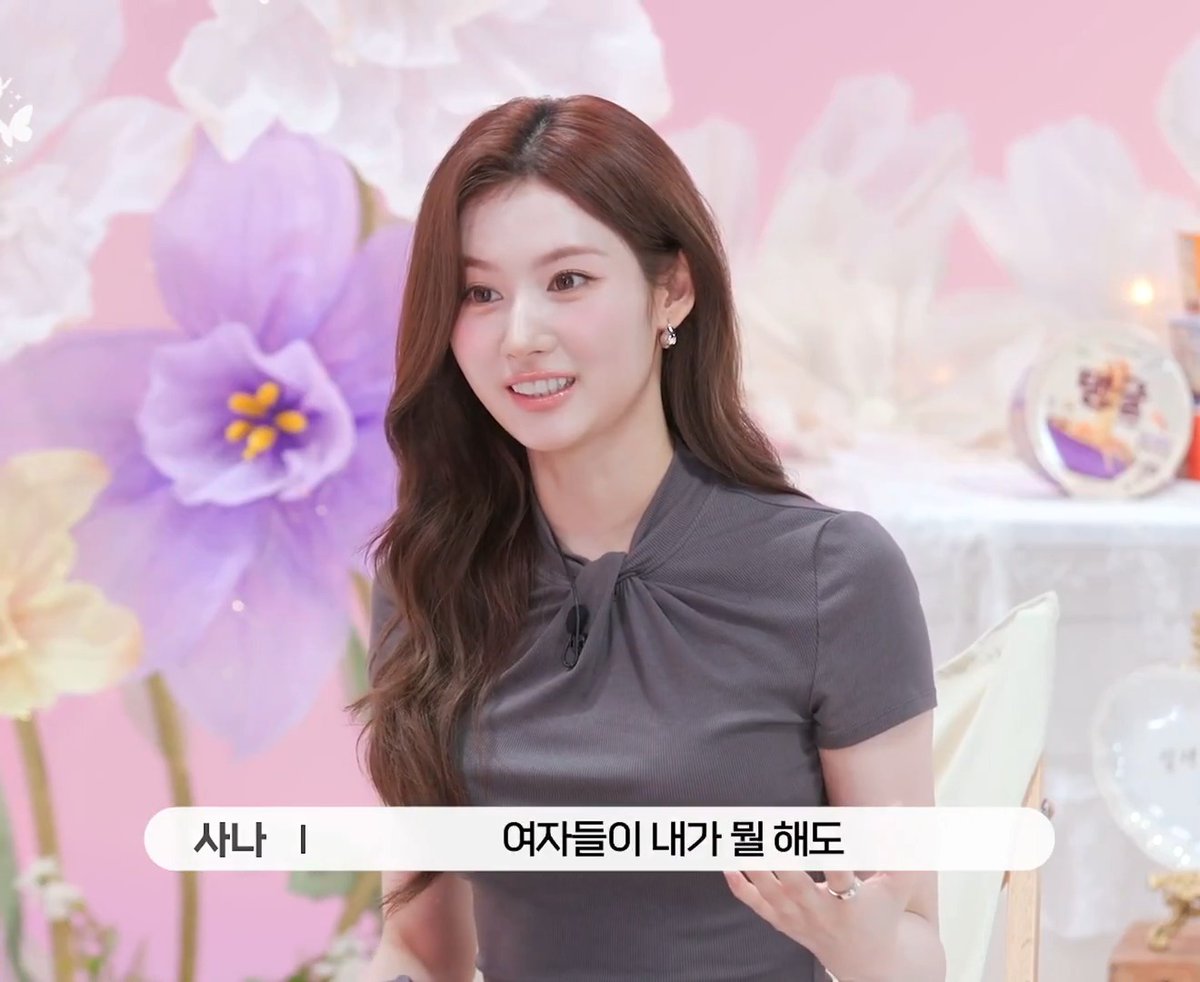 Sana, wearing a gray ruched top and drop earrings, smiles during what appears to be a variety show appearance against a floral pink set. The Korean subtitle reads "여자들이 내가 뭘 해도" (No matter what I do, women), referencing her playful comment about being jealous of how female fans react to male idols with screams.