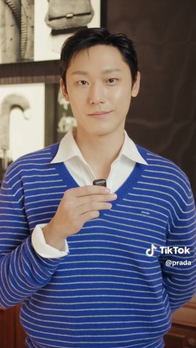 A young man with dark hair wearing a blue striped Prada sweater over a white collared shirt holds a small object, appearing in a TikTok video from '@prada'.