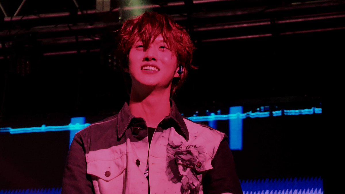 A young man with reddish hair smiles warmly on a dimly lit stage bathed in red and blue lighting, wearing a dark jacket over a lighter vest with 'EST. 1978' visible on it.