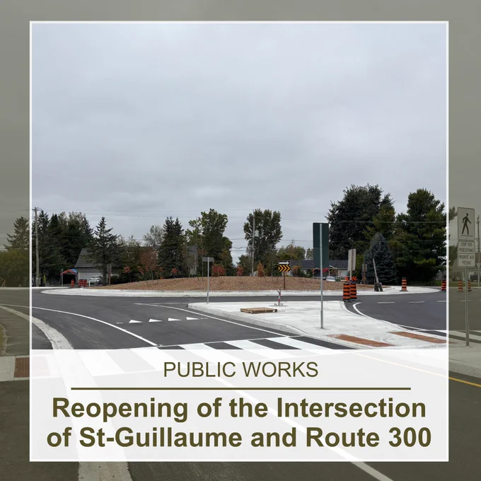 A roundabout with black and orange construction cones, trees, and two houses in the background, under a grey, cloudy sky; the text overlaid at the bottom center reads: "Public Works, Reopening of the Intersection of St-Guillaume and Route 300"