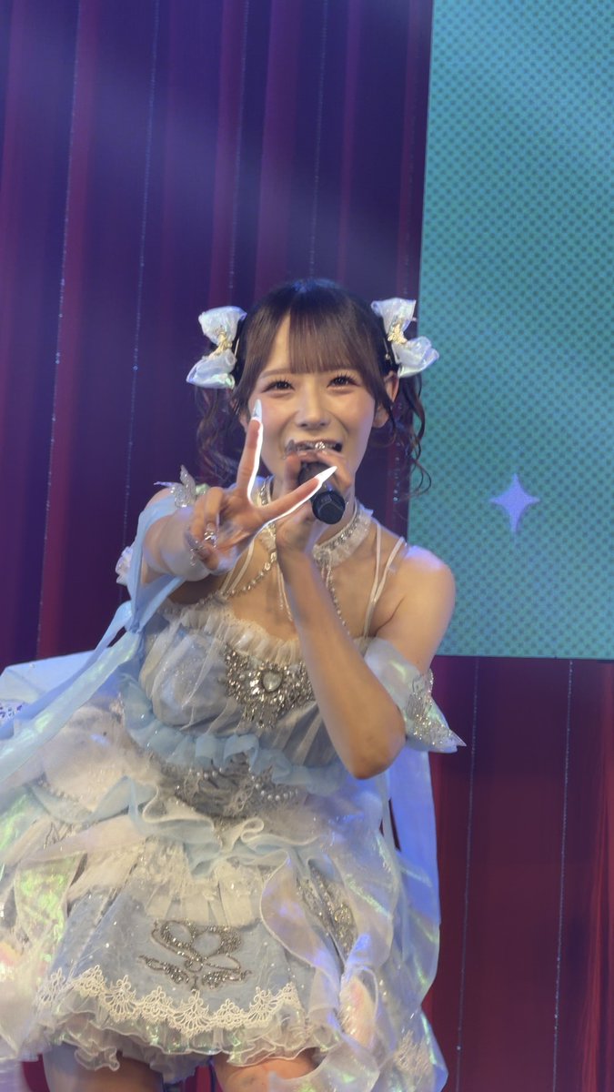Japanese idol performer on stage wearing an elaborate white and blue ruffled dress with bows, holding a microphone and making a peace sign.