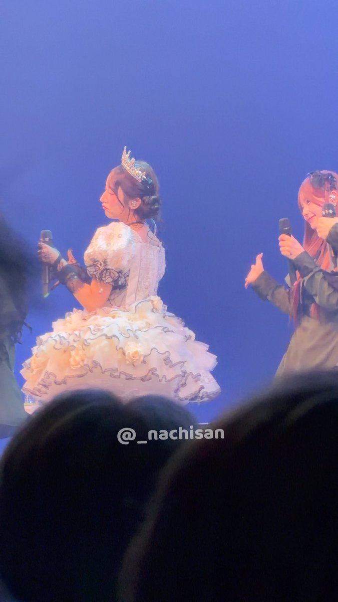 Two performers are on a blue-lit stage; one wears a princess-like white dress and crown, and the other wears a dark outfit with a red bow, with the watermark "@_nachisan" visible.