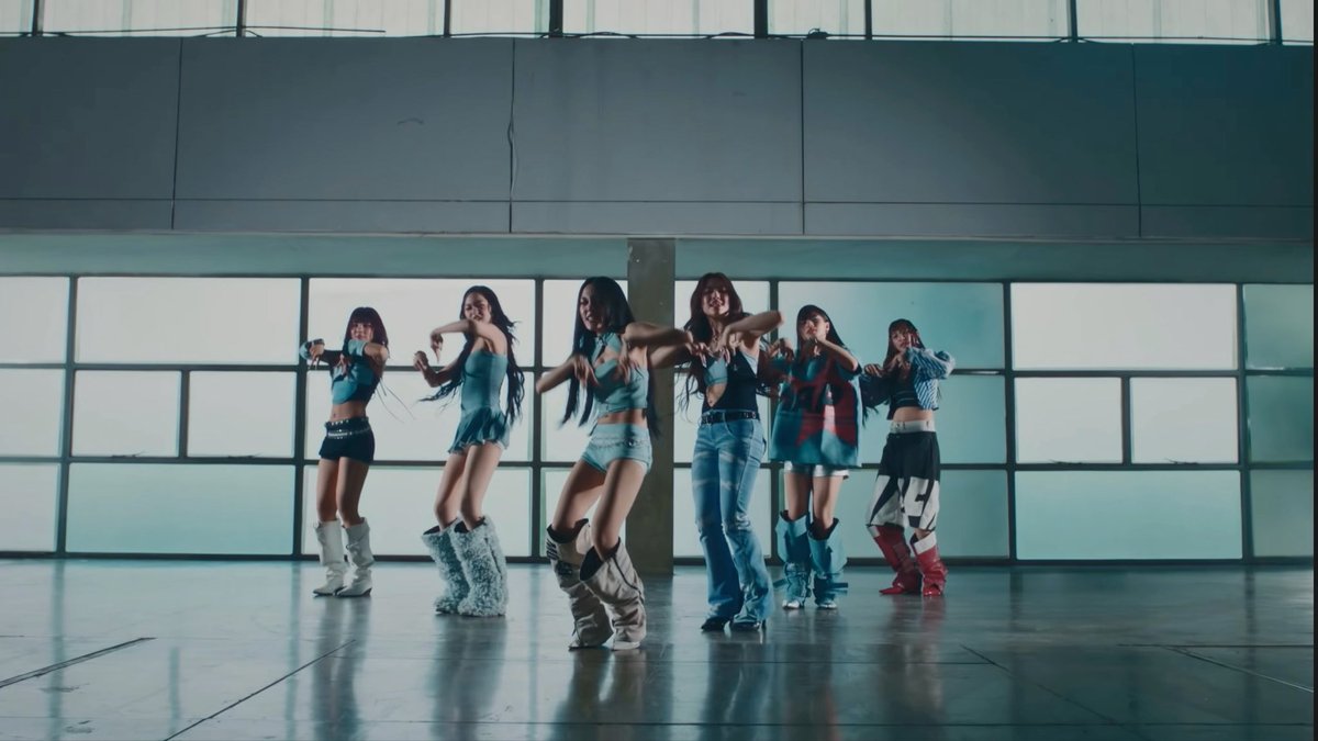 A group of six women in denim outfits and furry boots perform a choreographed dance routine in a spacious room with large windows.