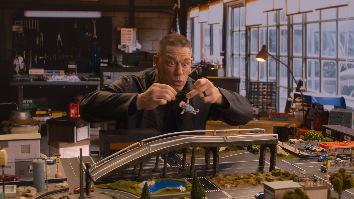 John Cena animatedly pretends to "rescue" a tiny figurine above an elaborate miniature cityscape featuring a bridge and detailed buildings. The scene ties into the playful, toy-centric nature of the upcoming Matchbox movie, where the Matchbox brand's iconic die-cast cars and worlds are brought to life on the big screen.