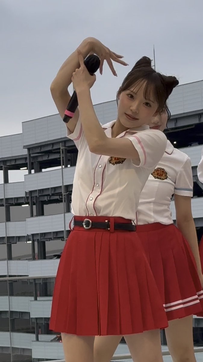 A young woman in a white blouse with pink piping and a red pleated skirt poses with a microphone, making a heart shape with her arms.