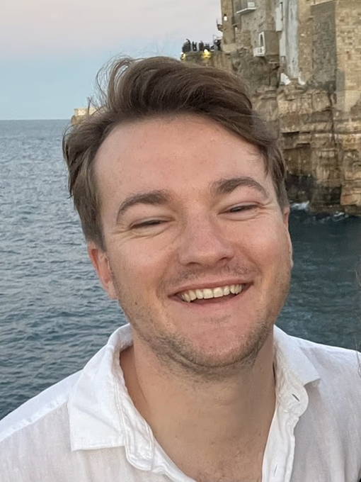 A young man with brown hair smiles broadly at the camera while wearing a white shirt, standing in front of a coastal cliffside and ocean. The image accompanies a post about Thomas Jewitt, presenting him as a cheerful, kind-hearted 32-year-old whose life was tragically cut short after being attacked at Notting Hill Carnival.