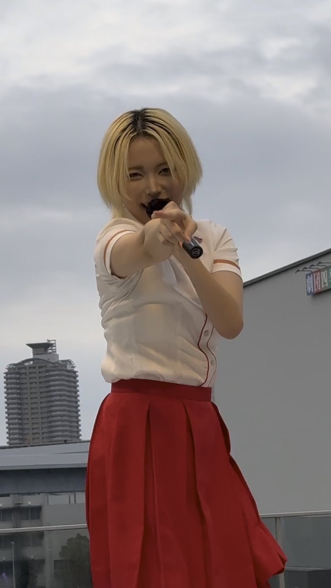 A blonde woman in a white button-up shirt and red pleated skirt holds a microphone and points directly at the camera while performing outdoors.