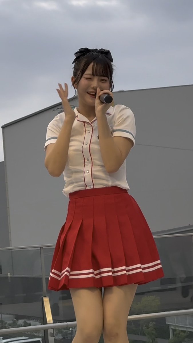 A young woman stands outdoors holding a microphone, wearing a white short-sleeved shirt with red piping and a red pleated cheerleader-style skirt with white stripes.