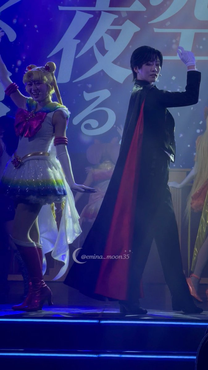 Two performers pose on stage in costumes resembling Sailor Moon characters, with a backdrop featuring large Japanese text and a watermark reading "@emina_moon35".